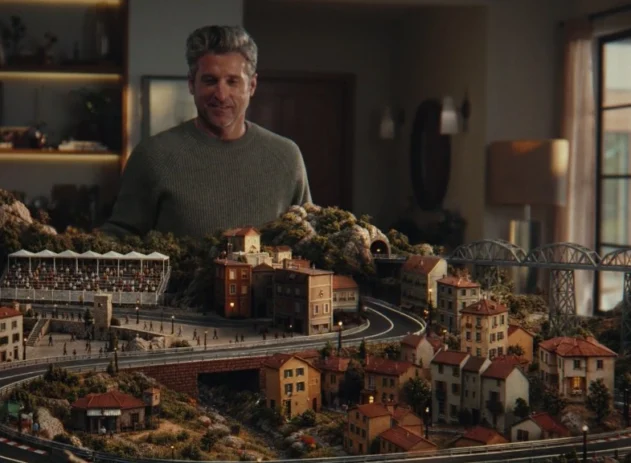 Patrick Dempsey aparece diante de uma pista de autorama em miniatura, cercada por casas, montanhas e arquibancadas, em cena da campanha do Nubank Ultravioleta.