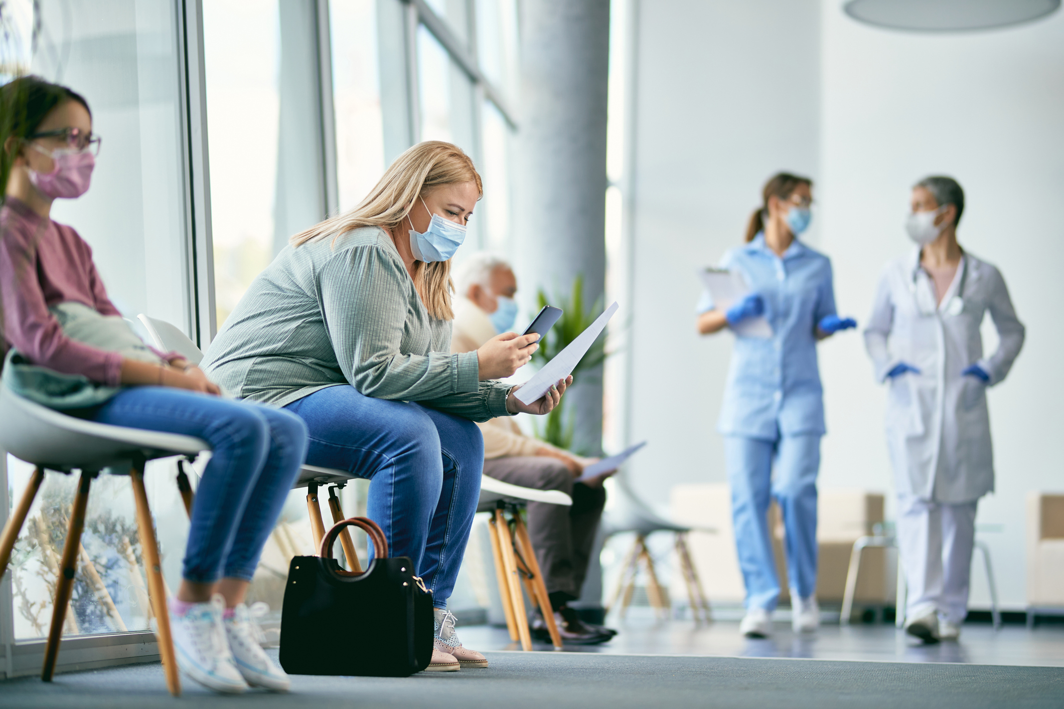 Frau mit Gesichtsmaske, die mit ihrem Smartphone ihren medizinischen Bericht im Wartezimmer der Klinik liest.