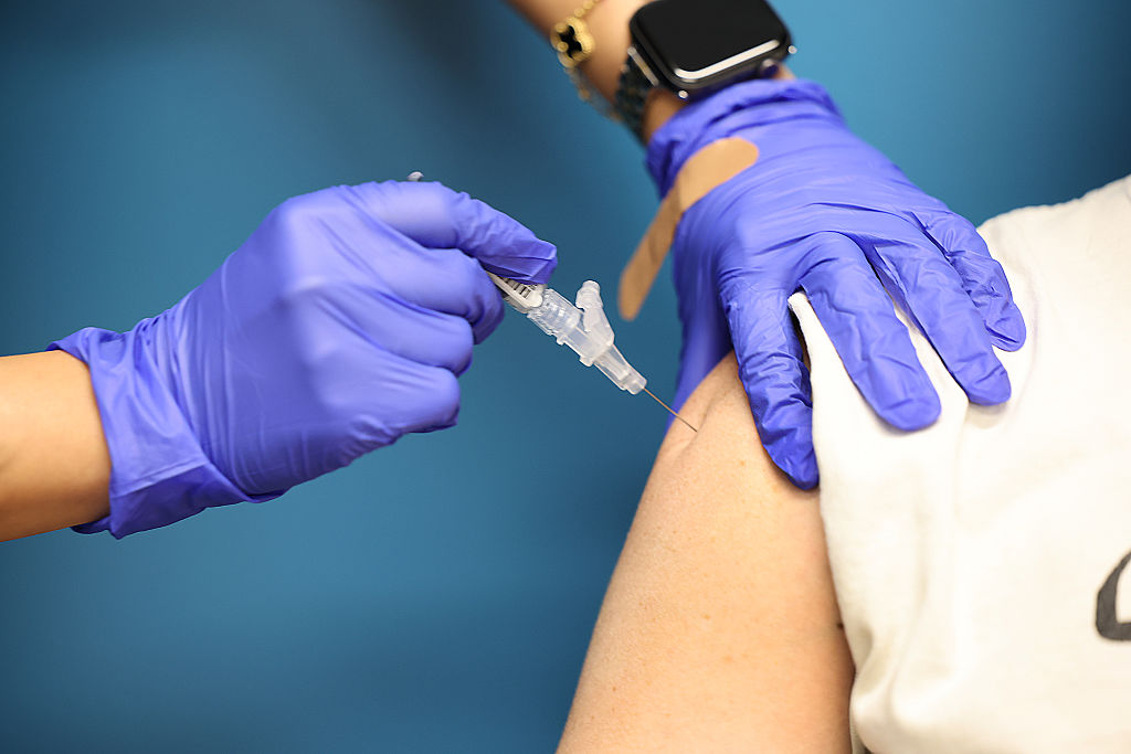 A medical professional with latex gloves inserts a vaccine into the shoulder of a patient with their sleeve rolled up. 