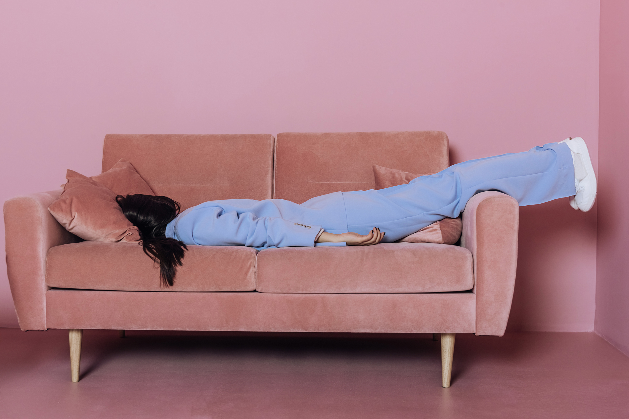 Seorang wanita sedang berbaring dengan baju warna biru di atas sofa berwarna merah muda. 