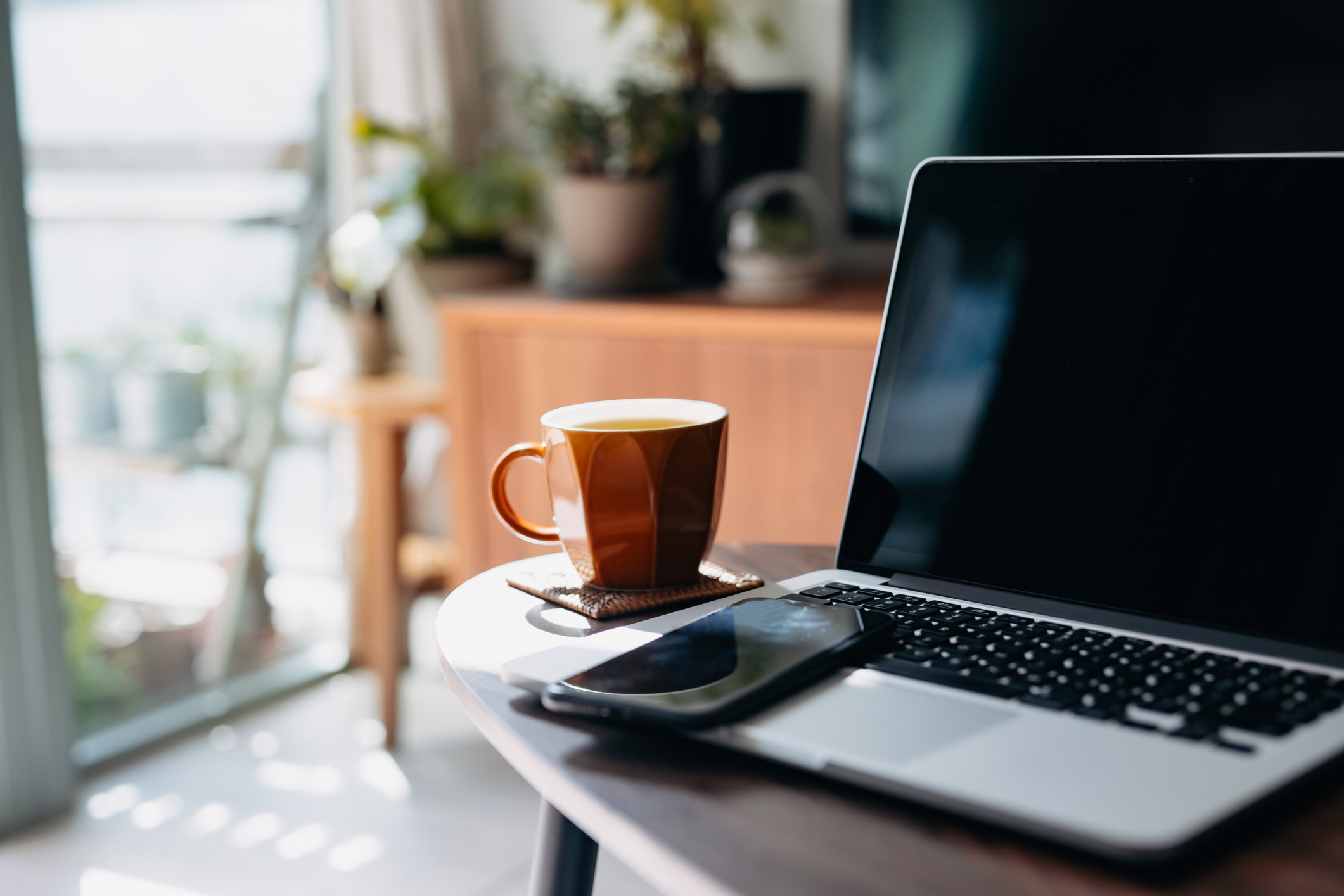 Gambar potongan meja kopi kayu dengan laptop, telefon pintar dan cawan teh di ruang tamu di rumah berhampiran tingkap menghadap cahaya matahari yang indah