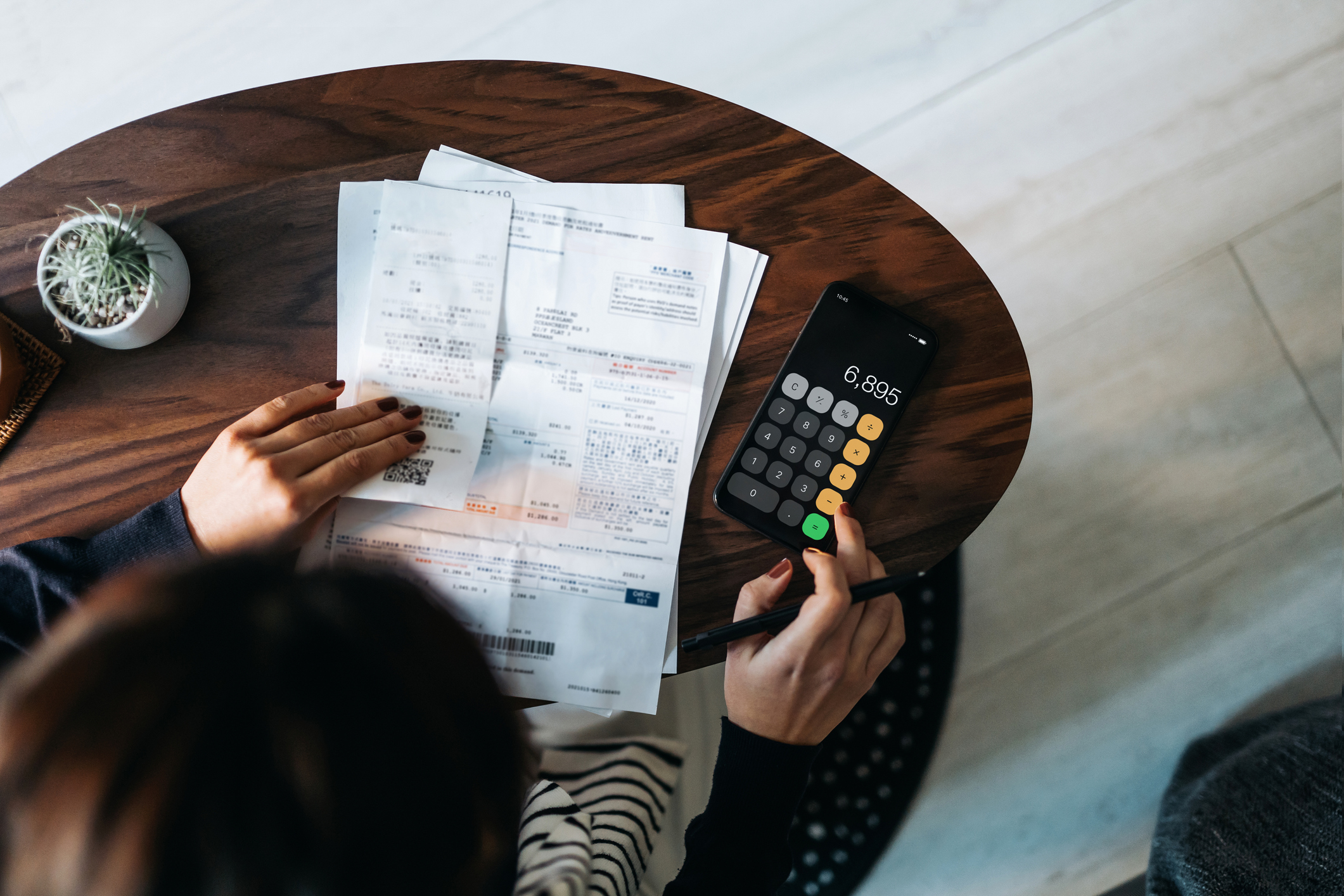 Overhead view of young Asian woman managing personal banking and finance at home. Planning budget and calculating expenses while checking her bills with calculator. Managing taxes and financial bills. Home budgeting. Concept of finance and economy