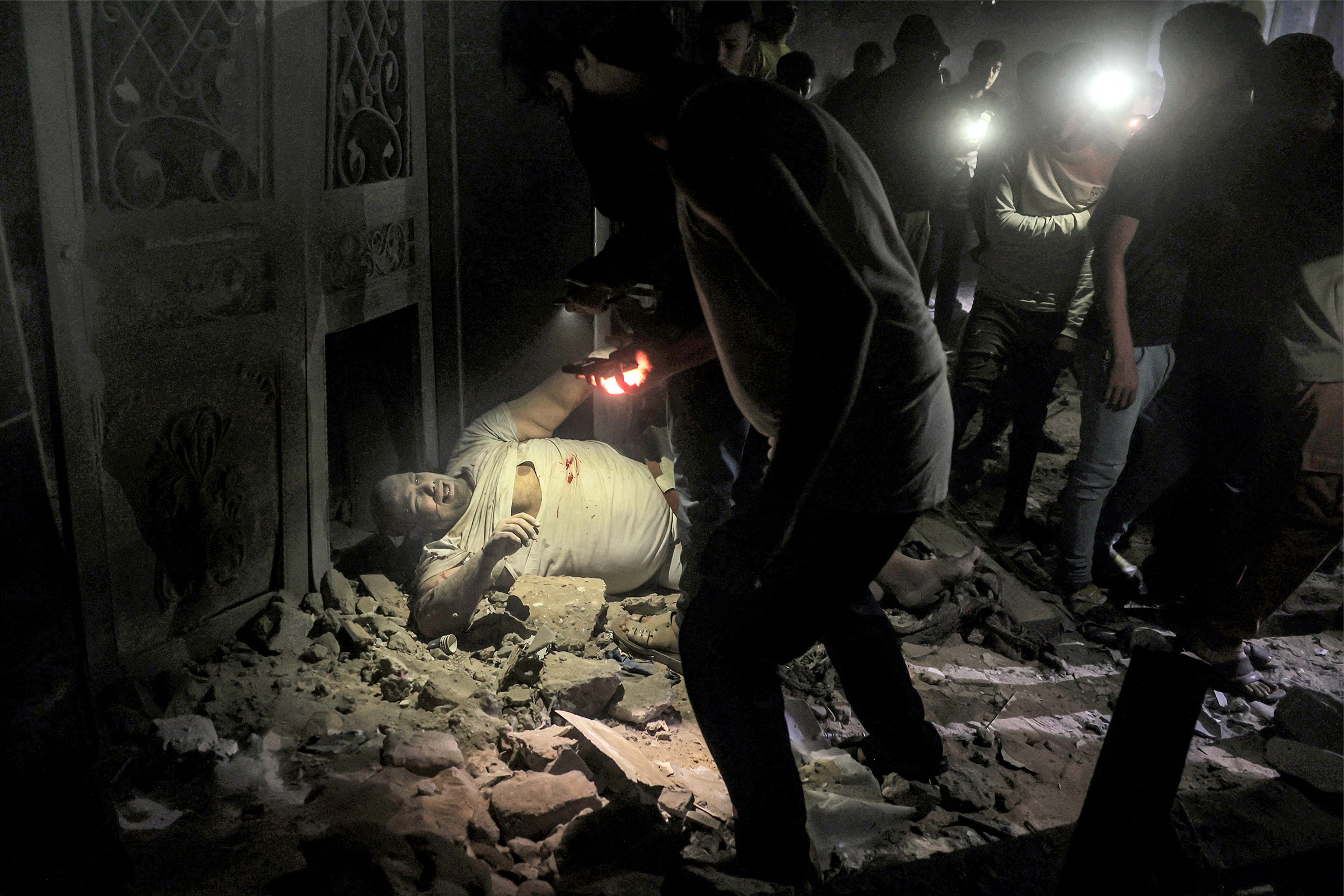拉法赫南部加沙地带一处建筑被以色列轰炸后,一名受伤人员躺在碎片中,周围人举着手电筒。