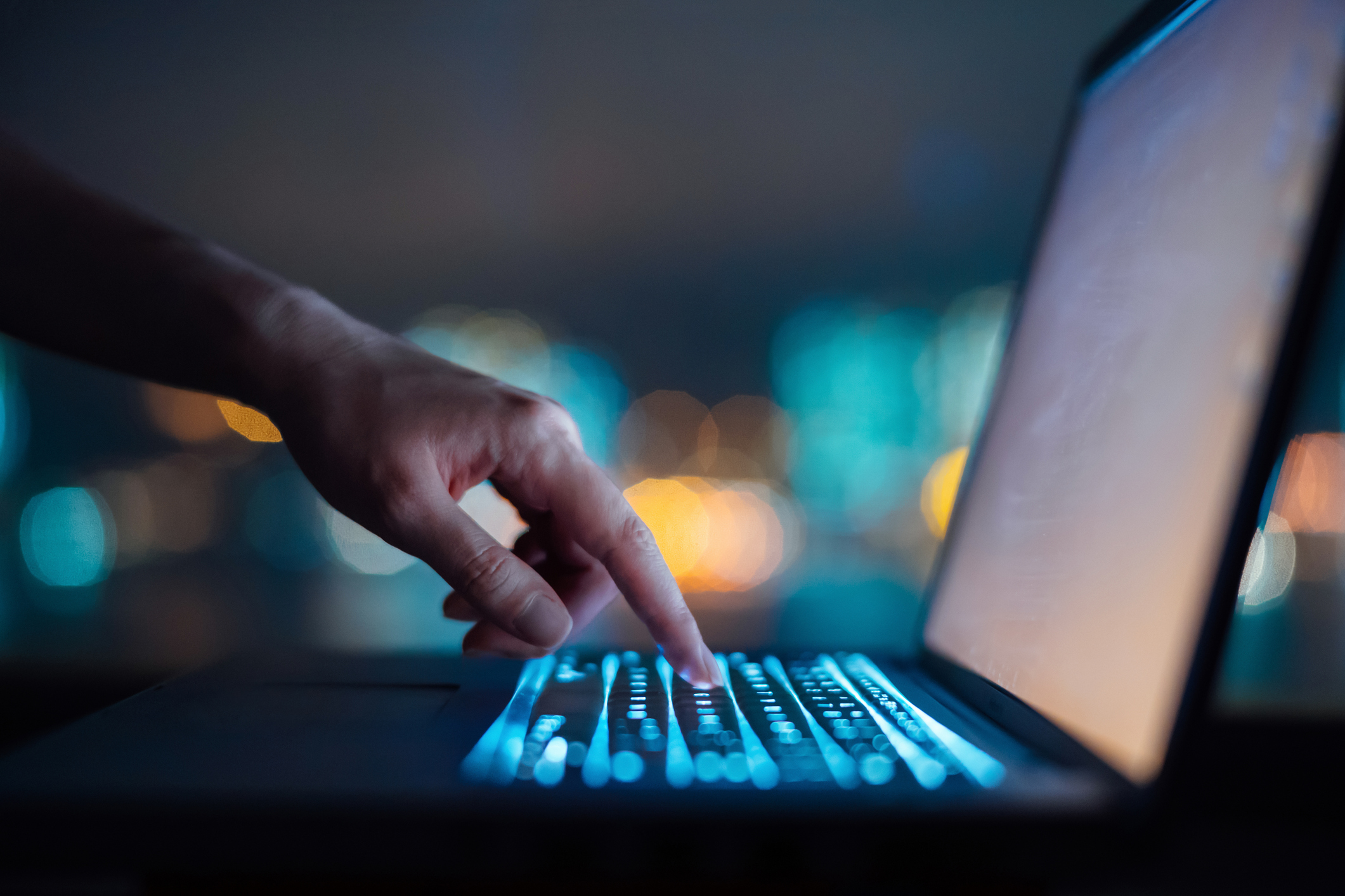 Close up of woman's hand typing on computer keyboard in the dark against colourful bokeh in background, working late on laptop at home