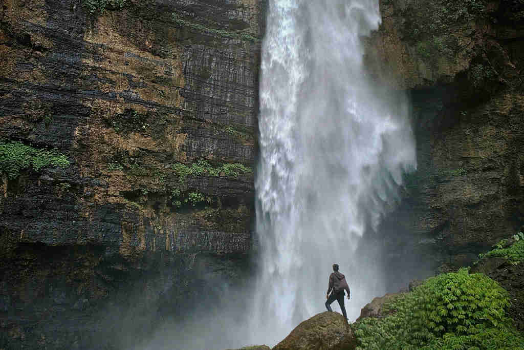 une cascade d'eau jaillit des rochers pour tomber en contrebas ou un admirateur est debout et admire la chute d’eau