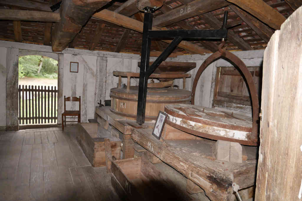 C’est une photo d’un ancien moulin en bois dans un cadre rustique. Le moulin est équipé d’une meule habillée de bois. Sur la droite, il y a une autre meule entourée d’un cerclage en fer et suspendue avec un outil pour la déplacer. La pièce a des murs et un plafond en bois, une fenêtre sur le côté droit et un banc en bois sur le côté gauche. Le sol est en bois et il y a une balustrade en bois à l’arrière-plan