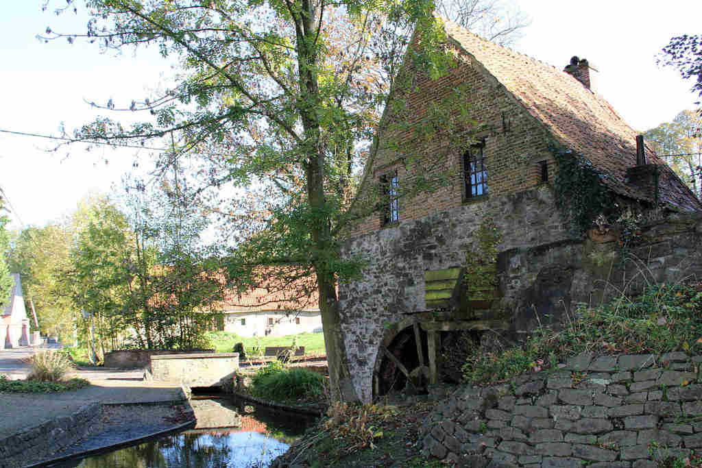 Maison en brique avec une roue à eau sur le côté, située à côté d’un canal et entourée d’arbres.