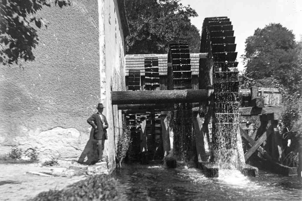 Cette image représente une photo monochrome de quatre énormes roues à aubes et une personne adossée au mur d’un énorme bâtiment qui doit être le moulin sur un fond de paysage d’arbres