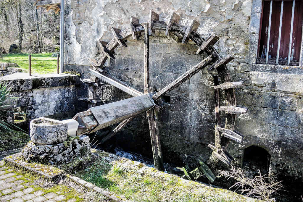 La photo présente une petite roue à aubes, située au cœur de l'ancien moulin de Bléfont. La roue est constituée de pales en bois fixées autour d'un axe central patinées de couleur grise par le temps