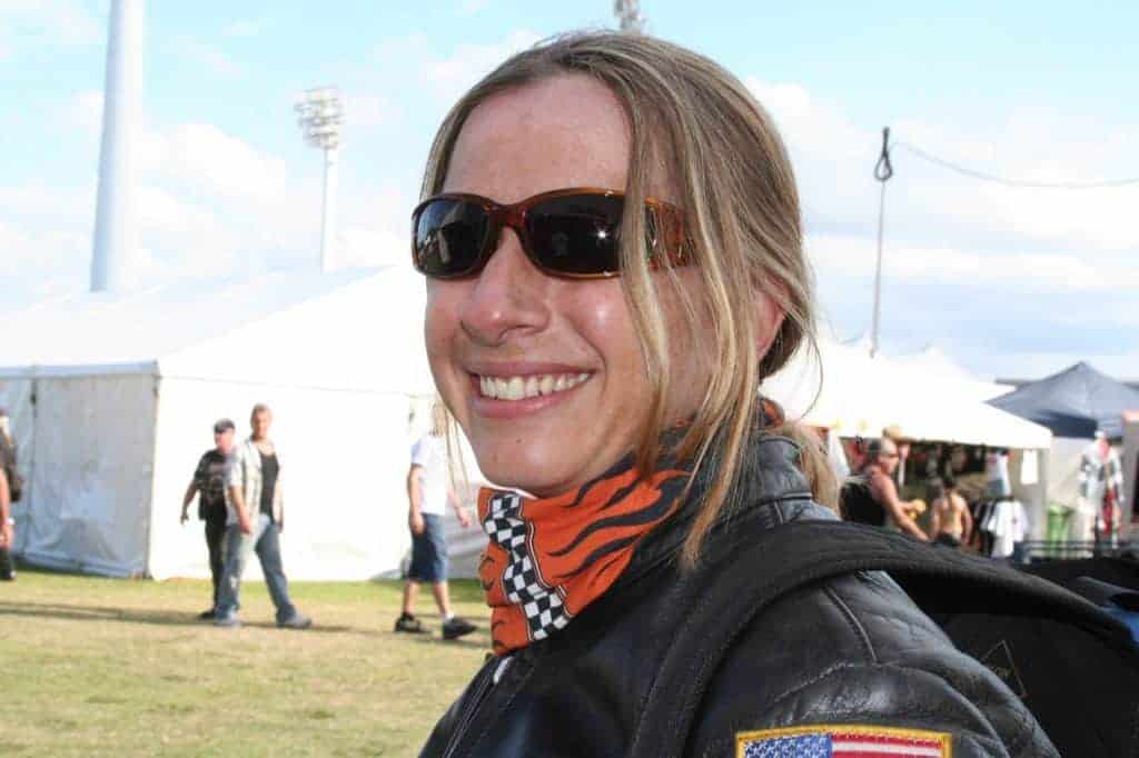 A side landscape shot of a woman in motorcycle leather gear. Kath is wearing a Original Buff® as scarf and smiling into the camera. I took the photo because I was amazed how straight and nice her hair was after having the helmet on for hours. She had the Original Buff® on as hair cover whilst wearing the helmet. The background shows the 2004 Gold Coast Bike Week with marquees and people leisurely walking around. Source: Edward Copyright: ©2004 Buff Downunder Pty Ltd