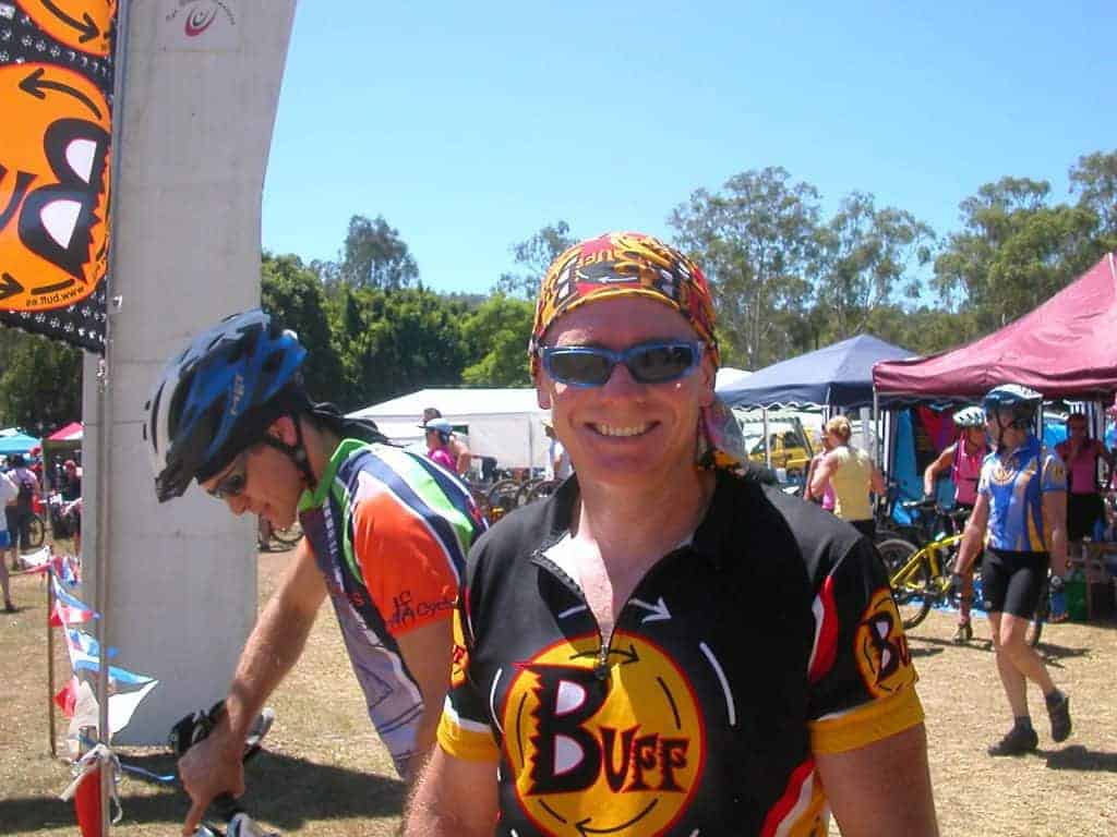 Landscape frontal shot photo of a male mountain bike rider. Scott smiling into the camera at the 2005 24 hour mtb race in Kooralbyne. It is hot. He is wearing a Original Buff® as helmet liner to cool the heat down. Source: Edward Copyright: ©2004 Buff Downunder Pty Ltd