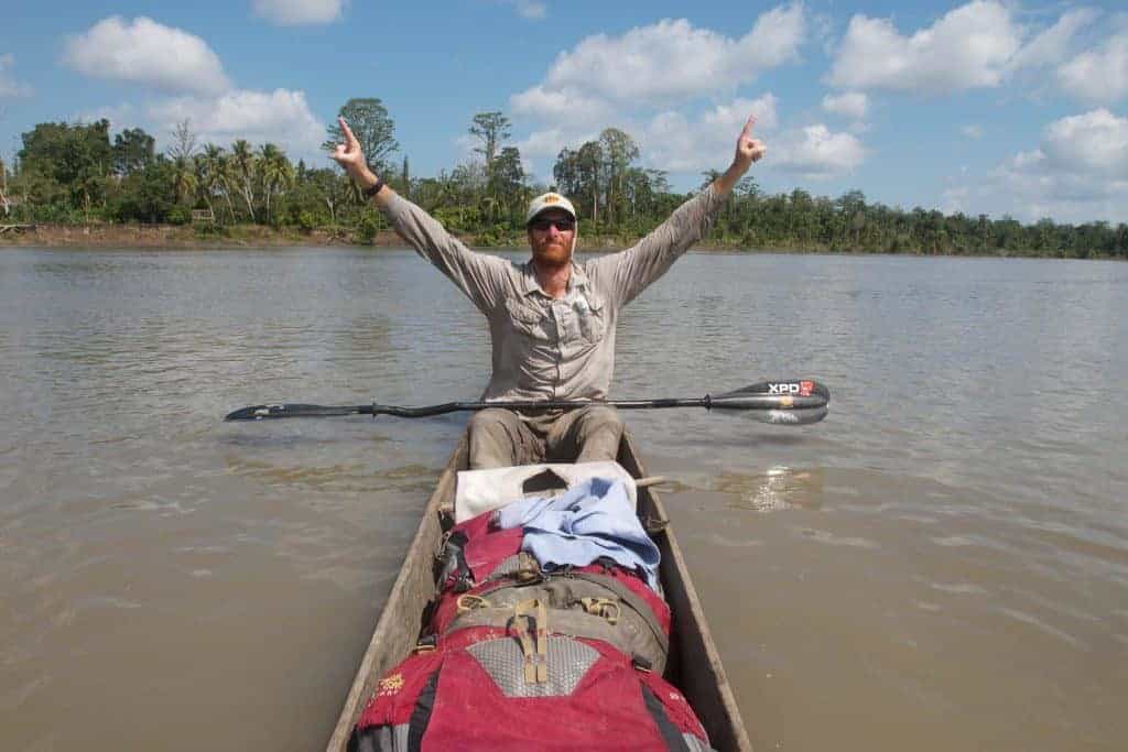 A frontal shot landscape photo of a man in a canoe raising his arms as a sign of victory. It is Andrew Johnson on the Sepik River in Papua New Guinea. He is wearing a Visor® Buff® as legionnaire style cap. The scene looks hot & humid. Image taken during the first spring to sea traverse of the Sepik River in Papua New Guinea. Source: Clark Carter © Clark Carter. http://www.adventureplaybook.com/