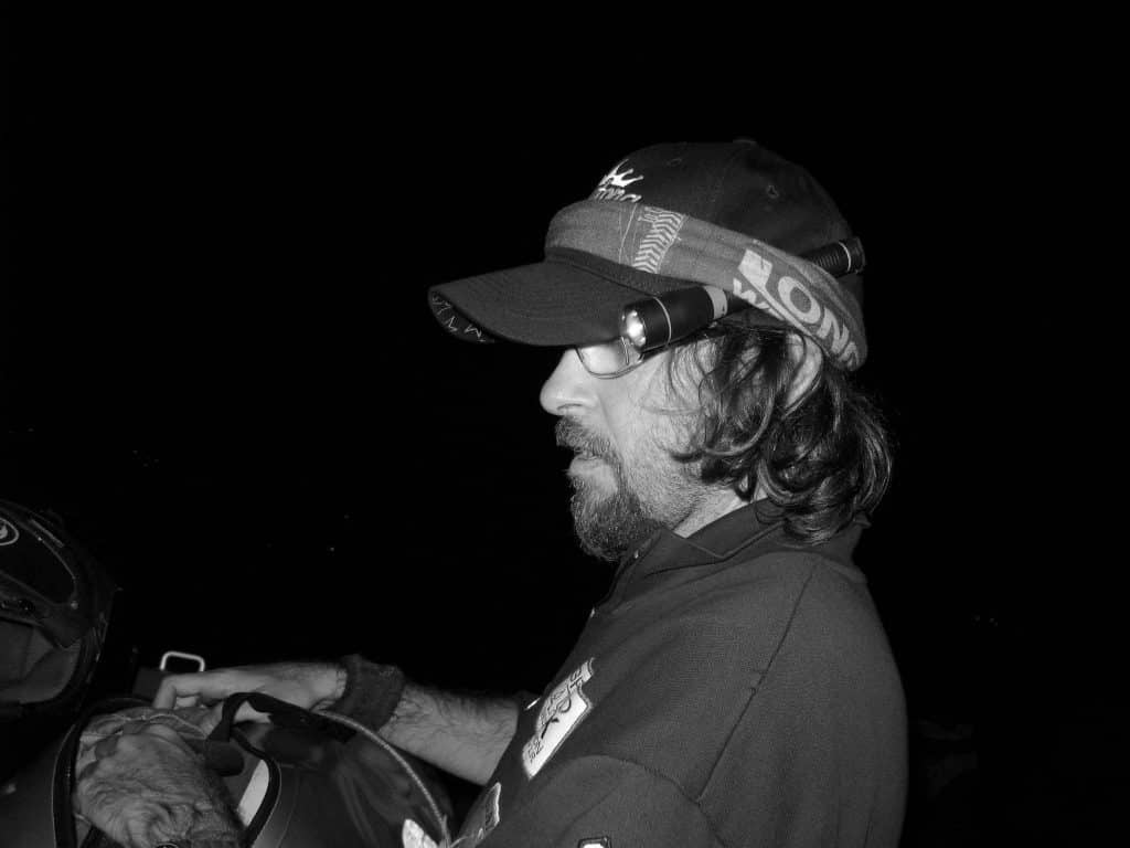 A side landscape shot of a man in the dark securing a hand torch with a Buff®. The man is wearing a polo shirt, a baseball cap and a Long Way Down Original Buff®. The Buff® is wrapped up tight around the cap and holds a hand torch in place. This way the man has both hands free for packing a bag. The photo was taken during a motorcycle tour so I assume they arrived their camp site in the the dark and have to set up camp in the dark. Source: buff.eu Copyright: Distributed for the promotion of the Original Buff® in motorcycle touring