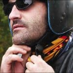 A side landscape shot of a man tying his motorcycle helmet. Visible is his upper body with the top of his helmet not visible. He is wearing a motorcycle leather jacket and a Original Buff® as scarf. His helmet is matt black and he is also wearing black sunglasses. He looks like a Harley rider. Interesting in this photo is how nice the Buff® closes up the jacket. It fills out all the nooks and crannies. The weather looks classic European summer. Not hot & not cold with a slight chill in the morning. Source: buff.eu Copyright: Distributed for the promotion of the Original Buff® in motorcycle