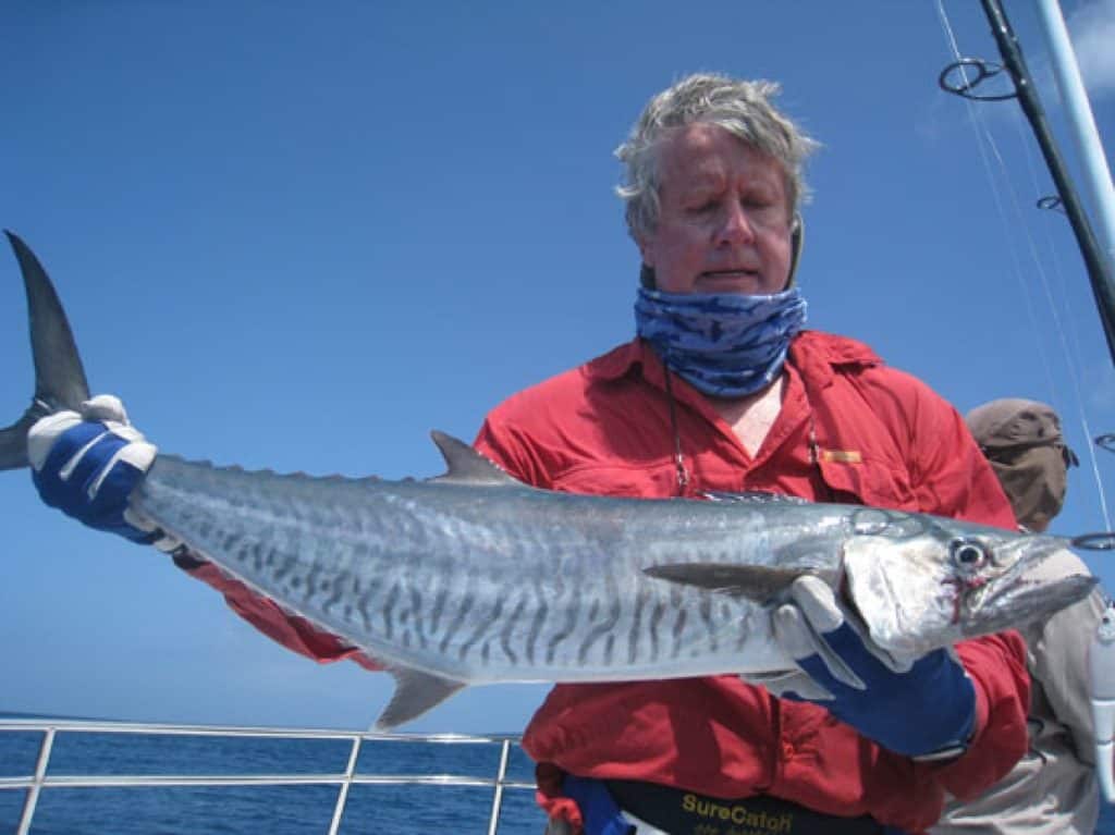 The landscape photo shows a middle aged man holding up his catch. He is wearing a Fishing High UV Buff® as scarf with the top covering his chin. The scenery looks like offshore fishing charter as the man wears tough leather gloves. Source: Unknown Copyright: Unknown