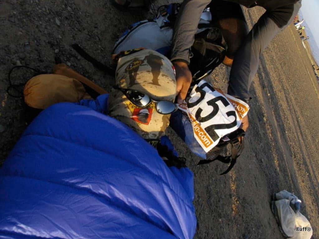 A landscape shot of a person lying on the ground in a sleeping bag. His entire head is covered with a Original Buff®. Another person is kneeling beside his head. He has put sunglasses on the sleeping persons face and is holding up the MDS event number 552. The photo was shot during the 2004 Marathon Des Sables. This is one way how to keept spiders and scorpions out during your sleep. Source: buff.eu © Distributed for the promotion of the Original Buff®