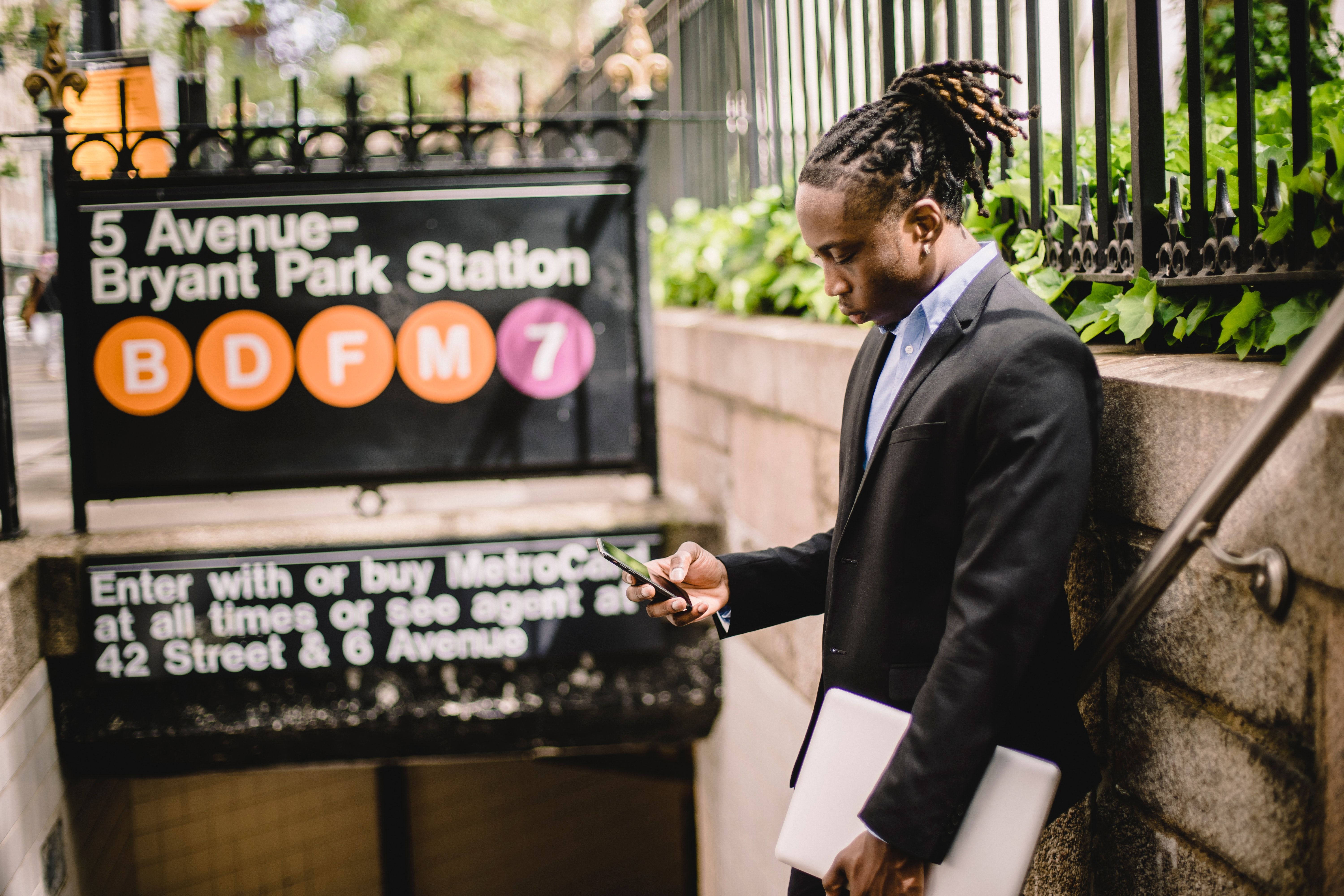 A man in a suit stands outside the entrance to the 5th Avenue Bryant Park Metro station in New York City, holding a closed laptop at his side and in his left hand, while he browses his smartphone with his right hand. 