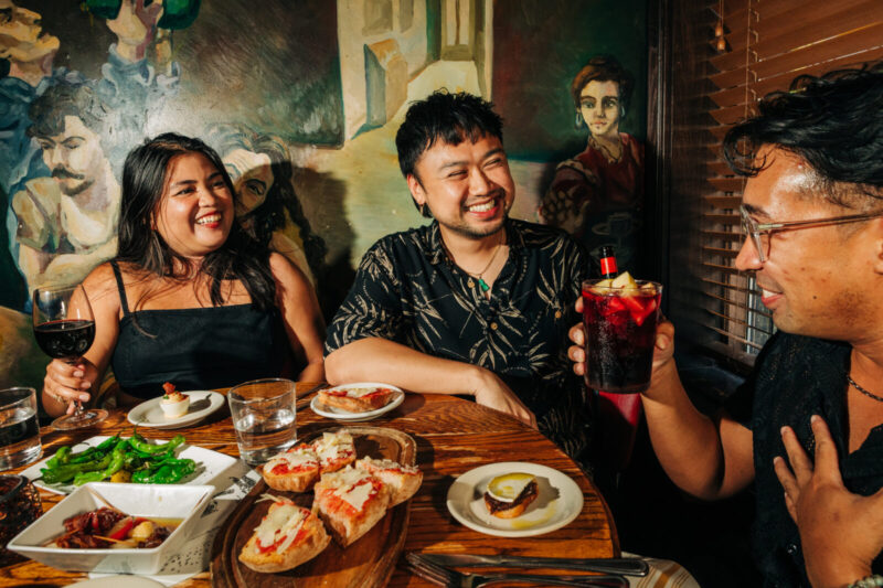 Three people enjoying Happy Hour dishes like shishito peppers alongside red wine and beer, available on Happy Hour.