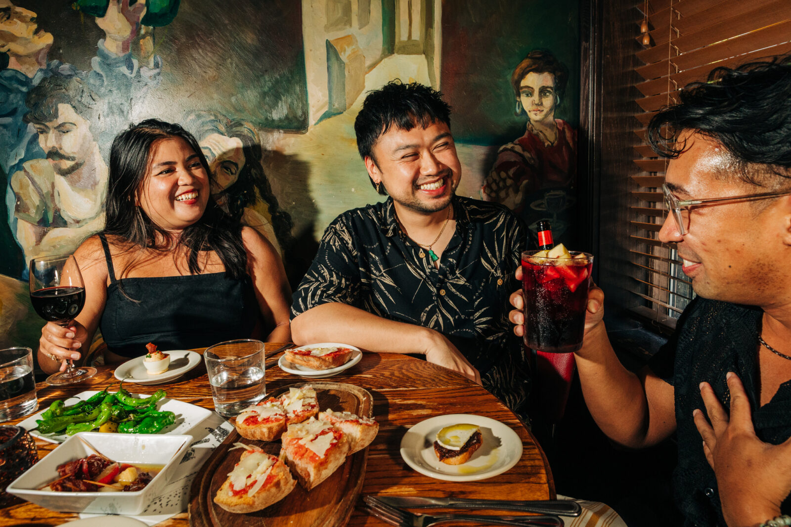 Three people enjoying Happy Hour dishes like shishito peppers alongside red wine and beer, available on Happy Hour.