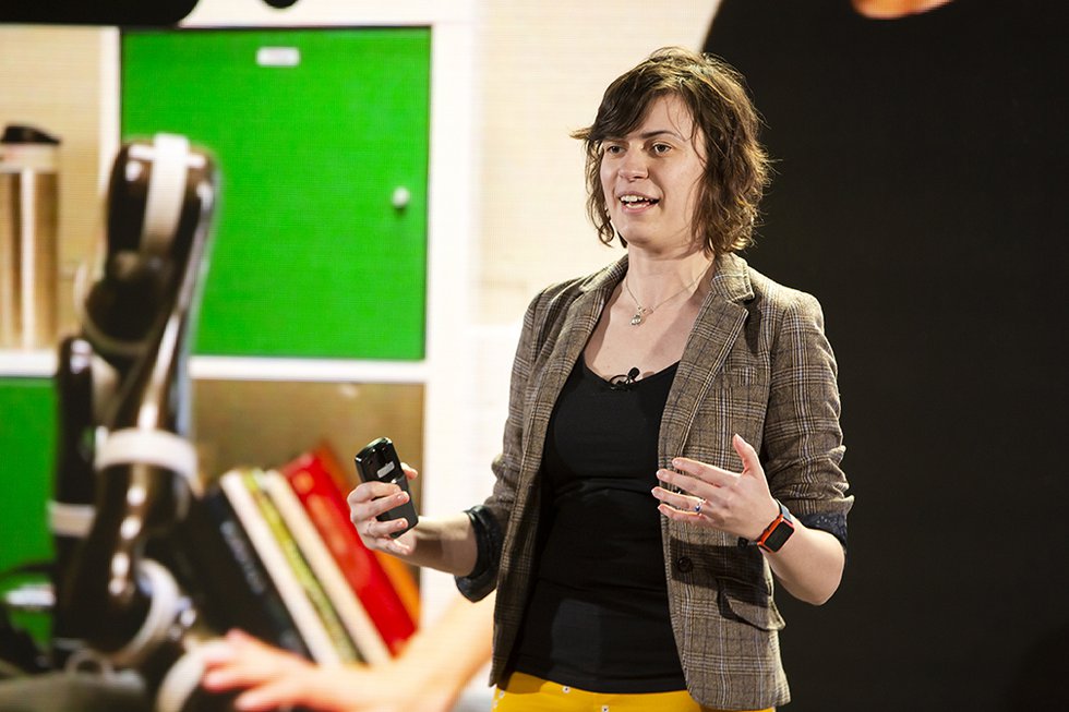 Photo of Anca Dragan onstage with a photo of a a robotics lab in the background