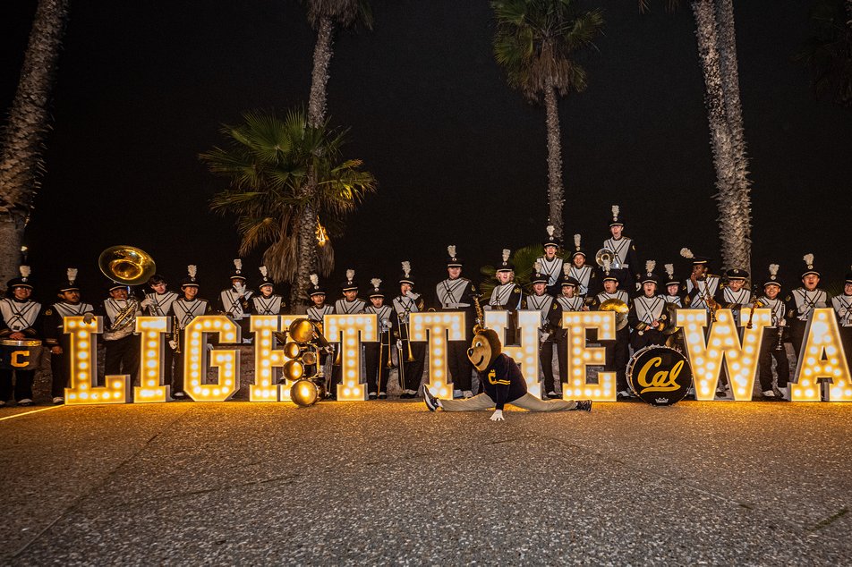 Color photo of students at night posing with giant light-up letters spelling "Light the Way" on a boardwalk.