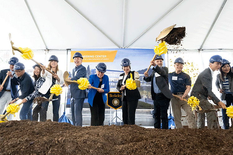 A group of people wearing hard hats and holding shovels with a yellow bow scoop the first bit of dirt.