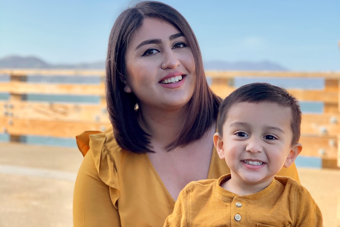 Photo of a young woman holding her toddler son under a blue sky, both smiling and wearing gold tops and blue jeans