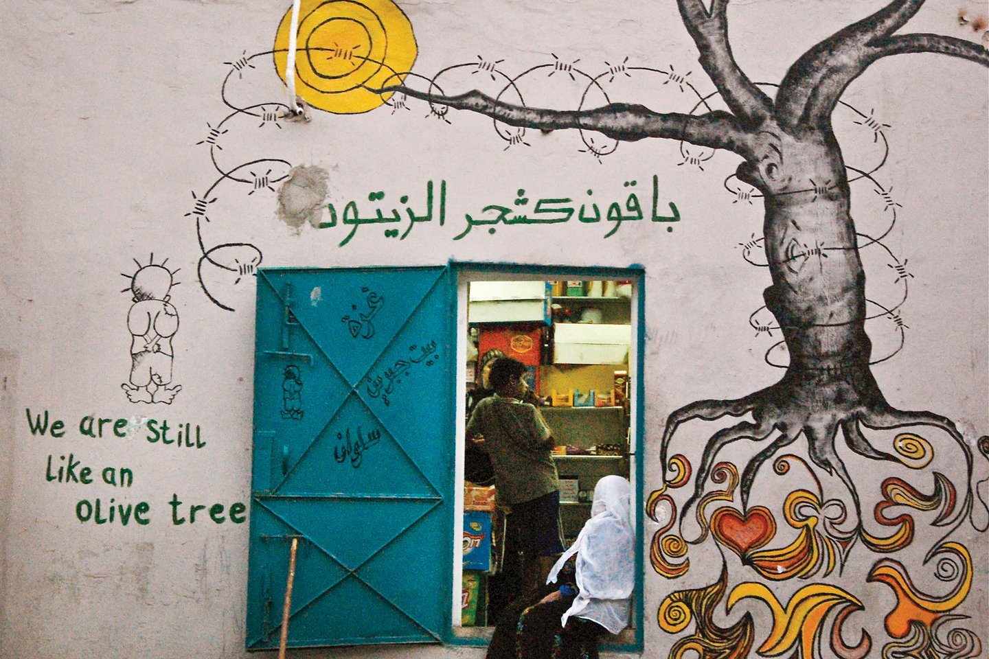 Photo of a shop with graffiti of a tree wrapped in barb wire and the words, "We are still like an olive tree."