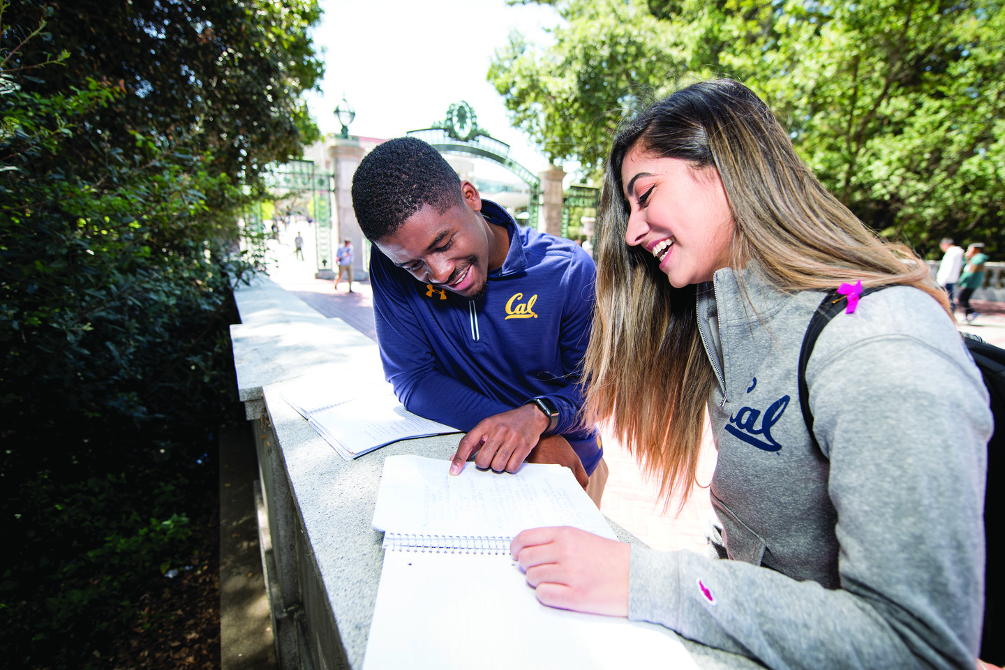 Color photo of two undergrads looking at assignments and smiling, Sather Gate visible in the background