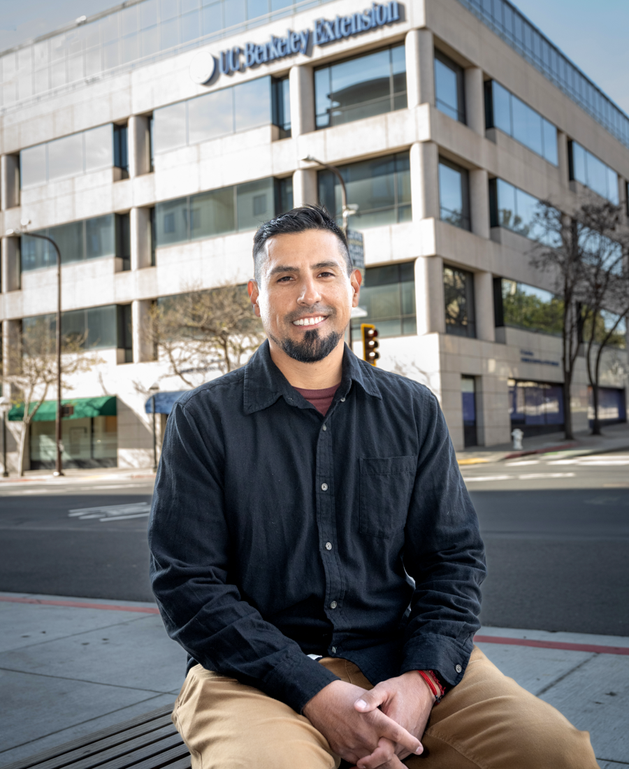 Juan is seated on a bench, with a black shirt and tan pants, and a building in the background.
