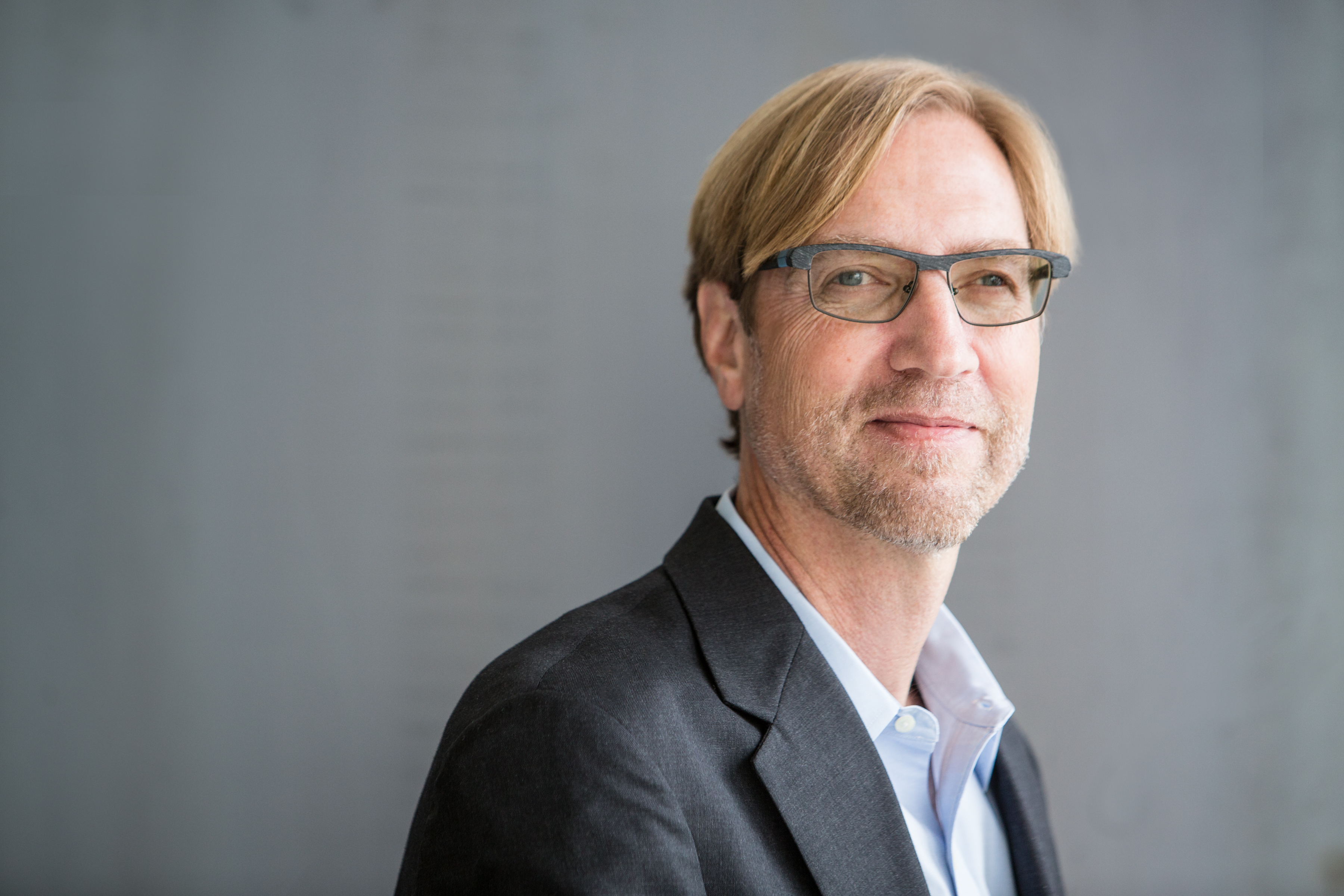 Color headshot of a blond man in a gray suit jacket and light-colored shirt, wearing glasses and smiling slightly.