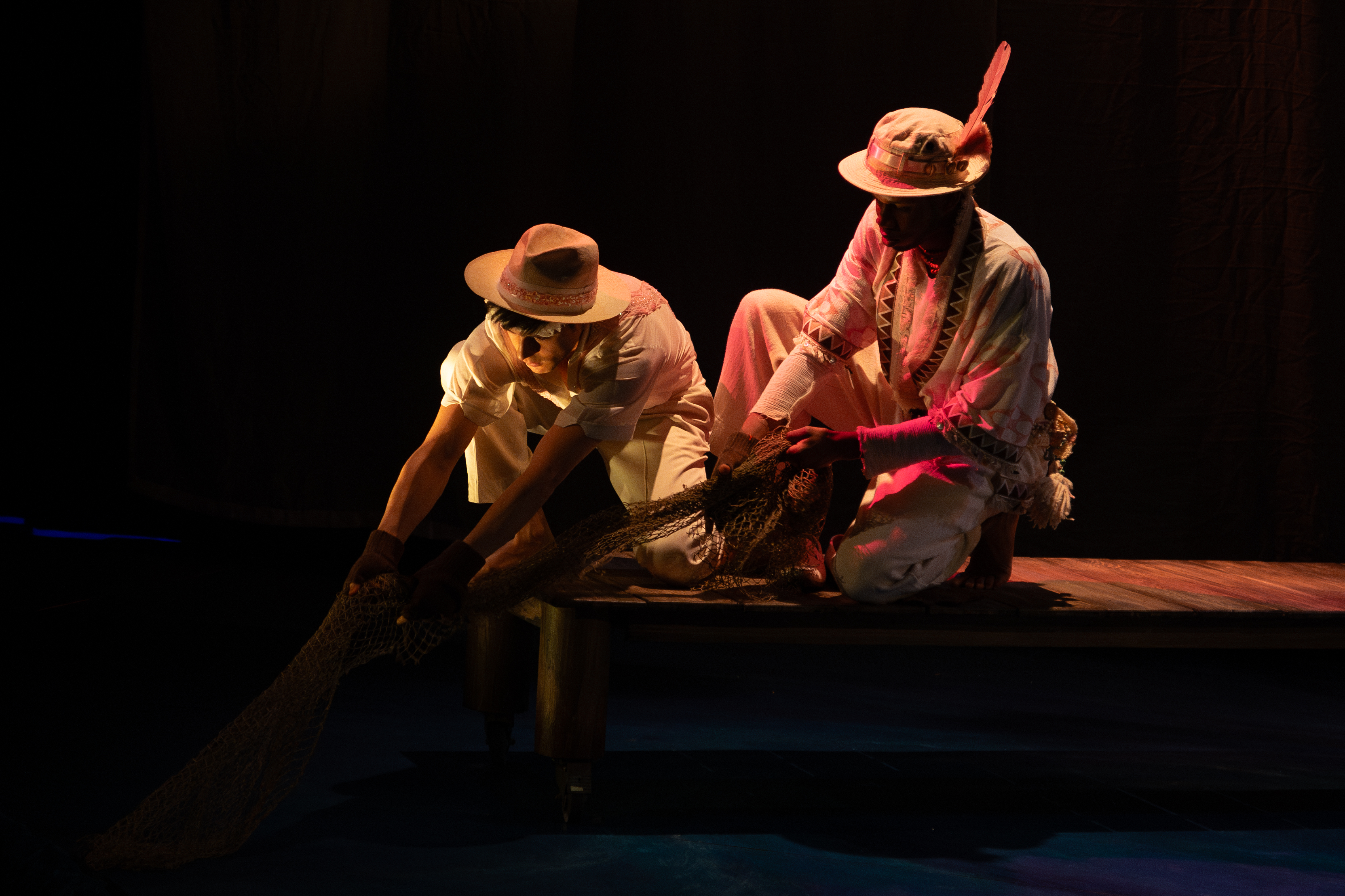 Photo of two students in traditional Brazilian clothing, kneeling on a set piece of a dock and pulling a fishing net from the water.