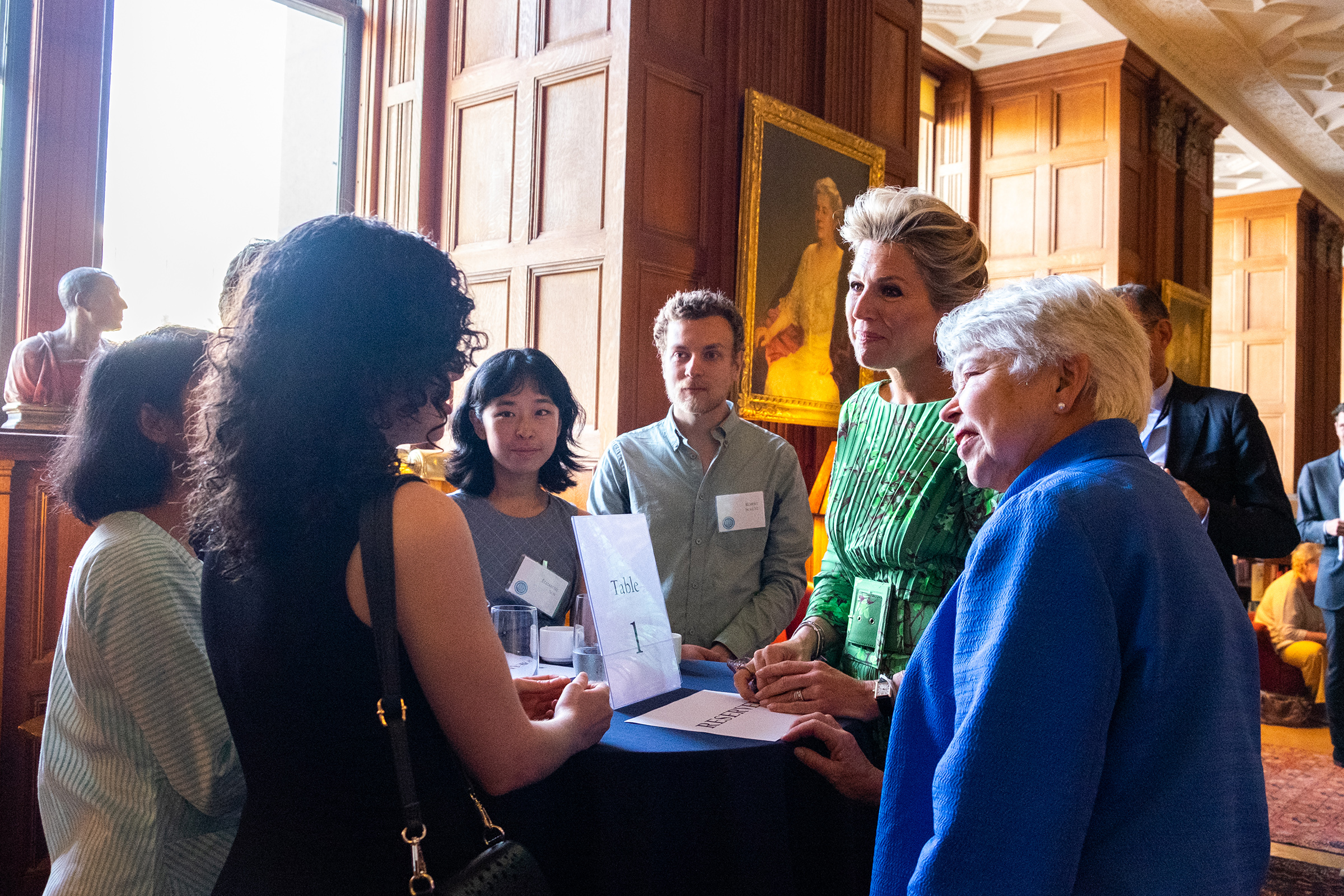 Photo of the chancellor, wearing a blue jacket, and the queen, wearing a green dress, engaged in lively conversation with students.