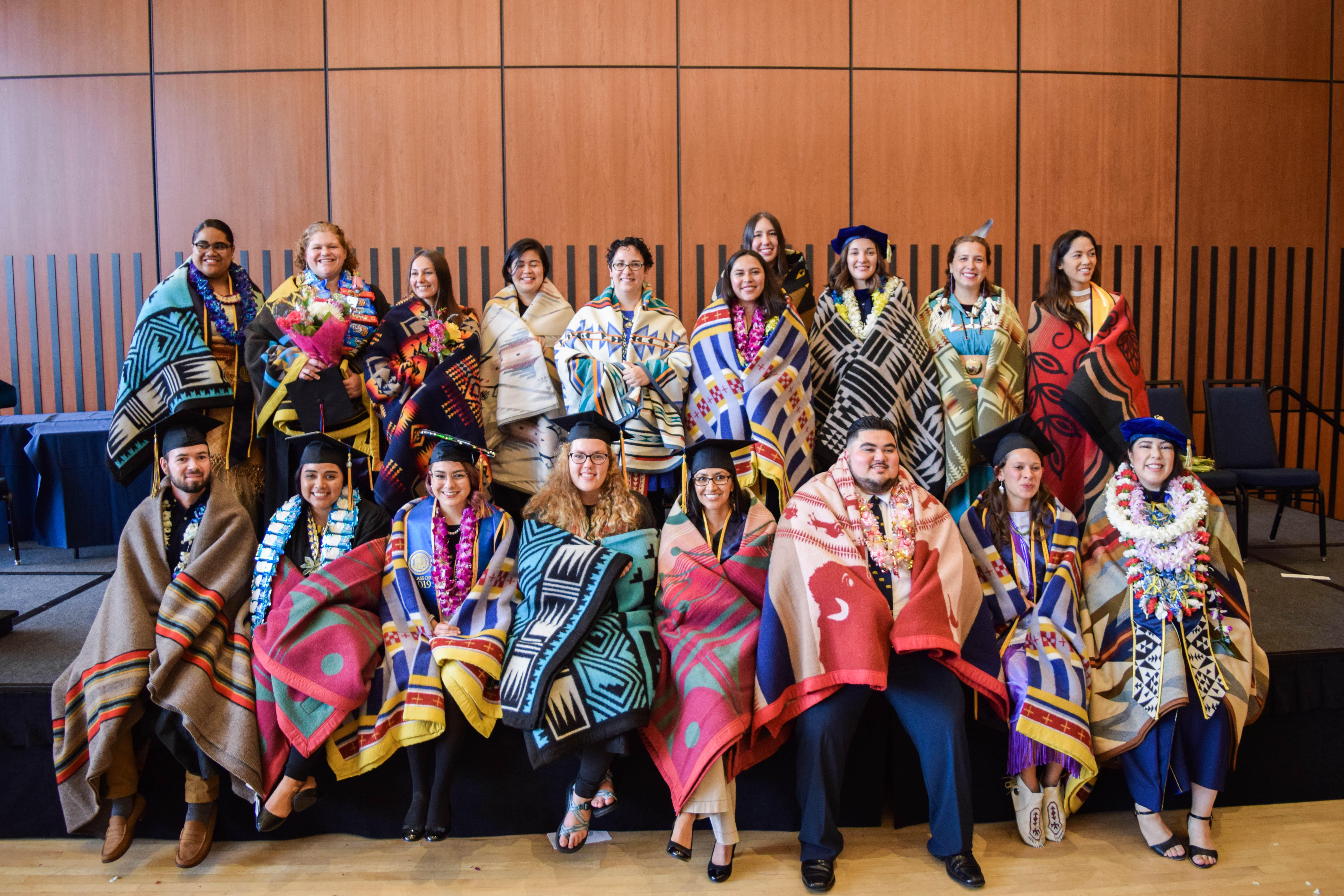 Photo of about 20 graduate students wearing graduation caps and wrapped in traditional Native American blankets.