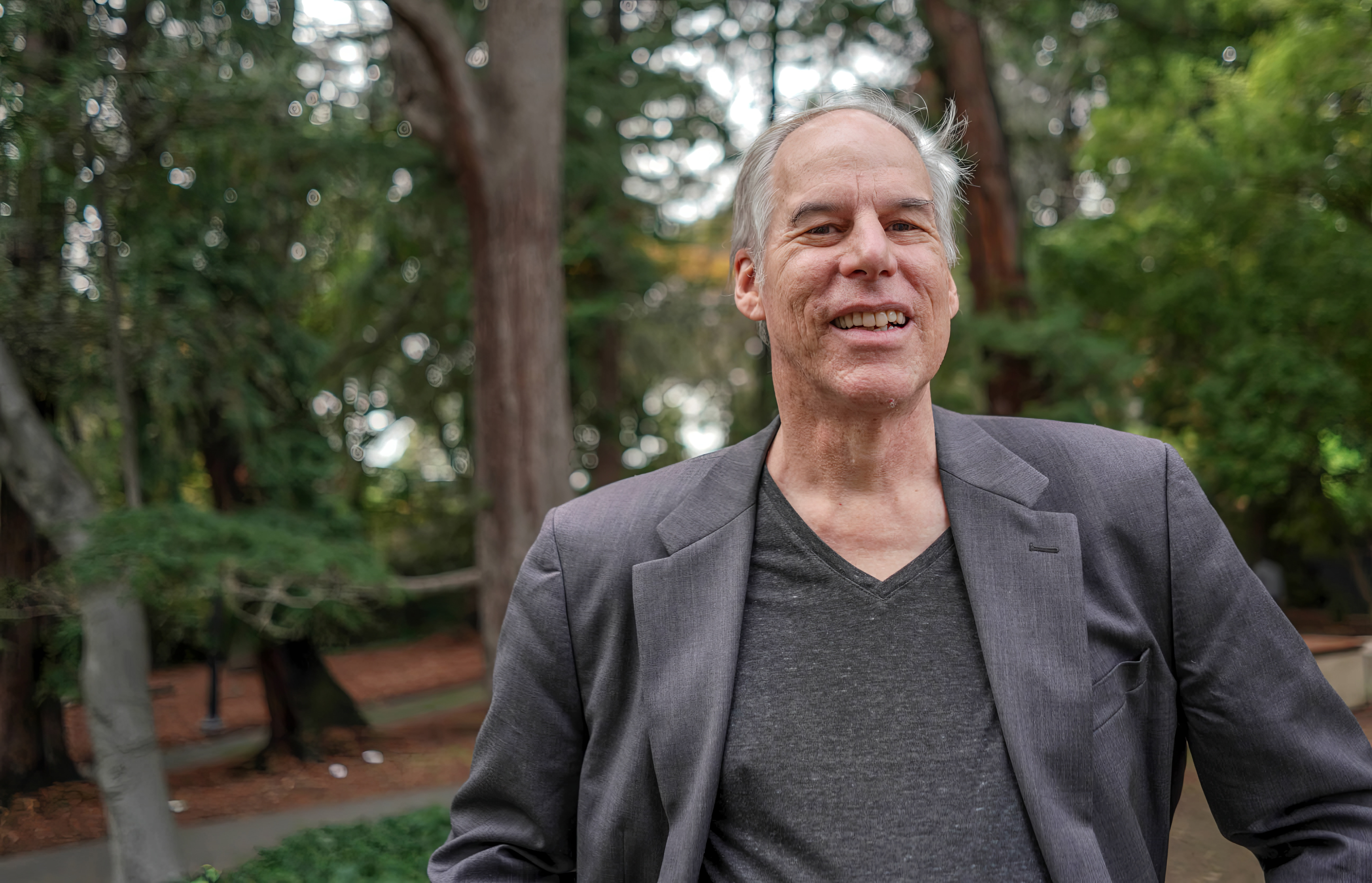 Photo of Alan wearing a gray top and jacket on campus in front of trees and paths.