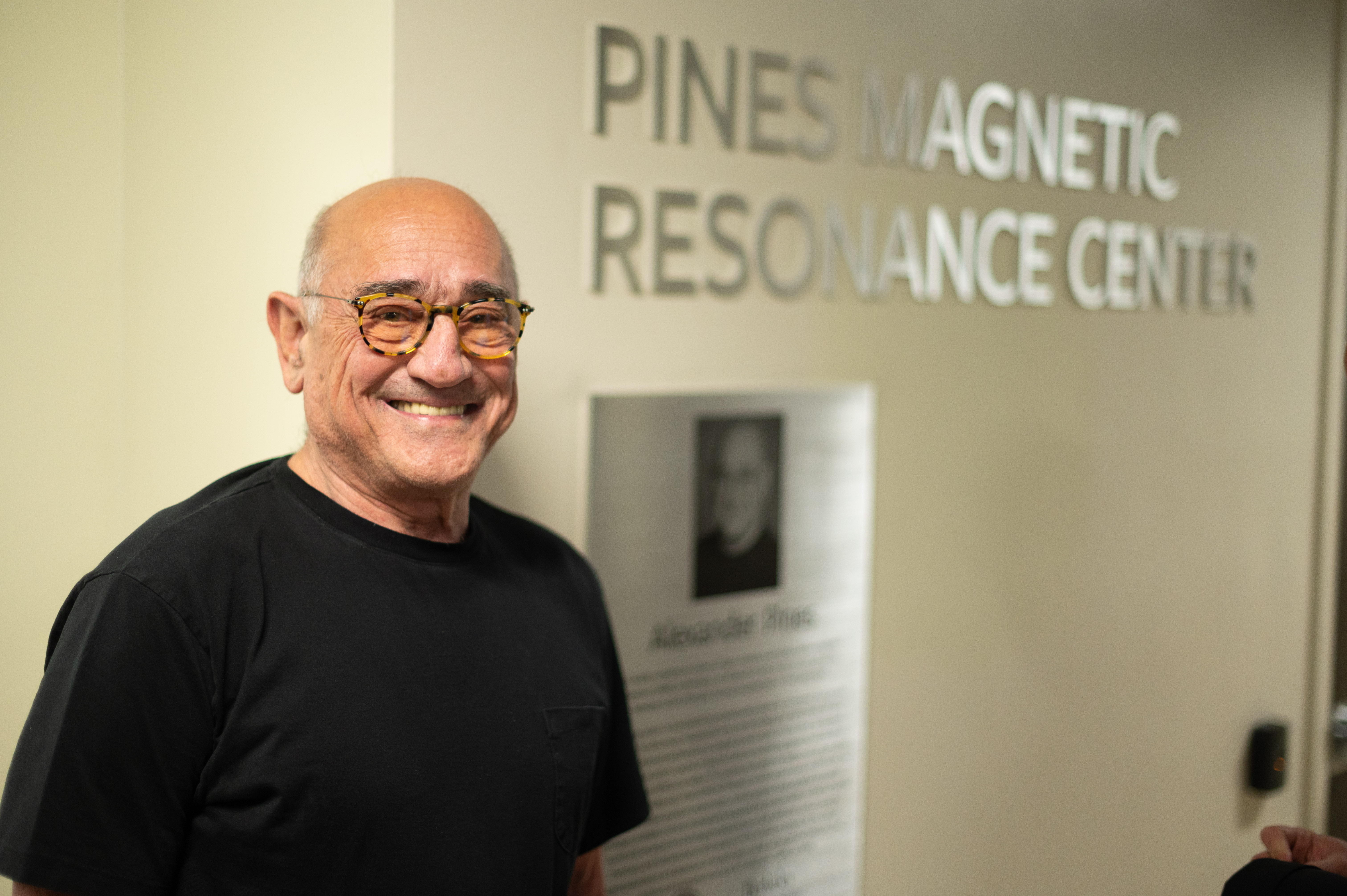 Photo of Alex in a black T-shirt and tortoise shell glasses, standing in front a new sign for the center.