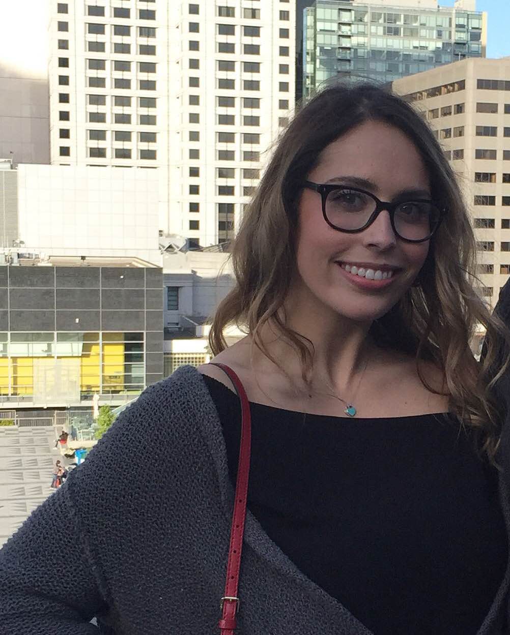 Color headshot photo of Alexis smiling at the camera, wearing glasses, high-rise buildings in the background