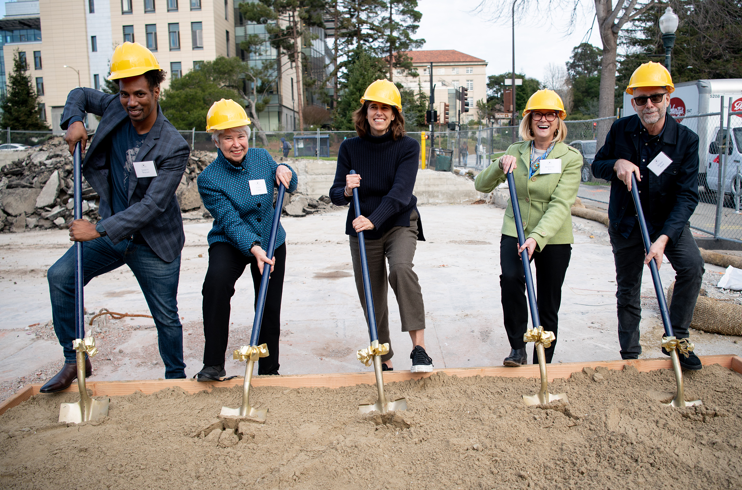 Photo of five people wearing bright yellow construction hats holding shovels, prepared to dig into dirt.