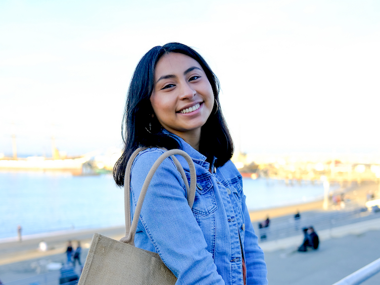 Photo of Angelica wearing a blue jean jacket and carrying a tan bag. She has shoulder-length black hair.