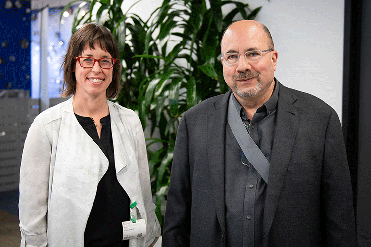 Photo of Ann in a black top and white jacket and Craig in a gray shirt and jacket. A large green plant is behind them.