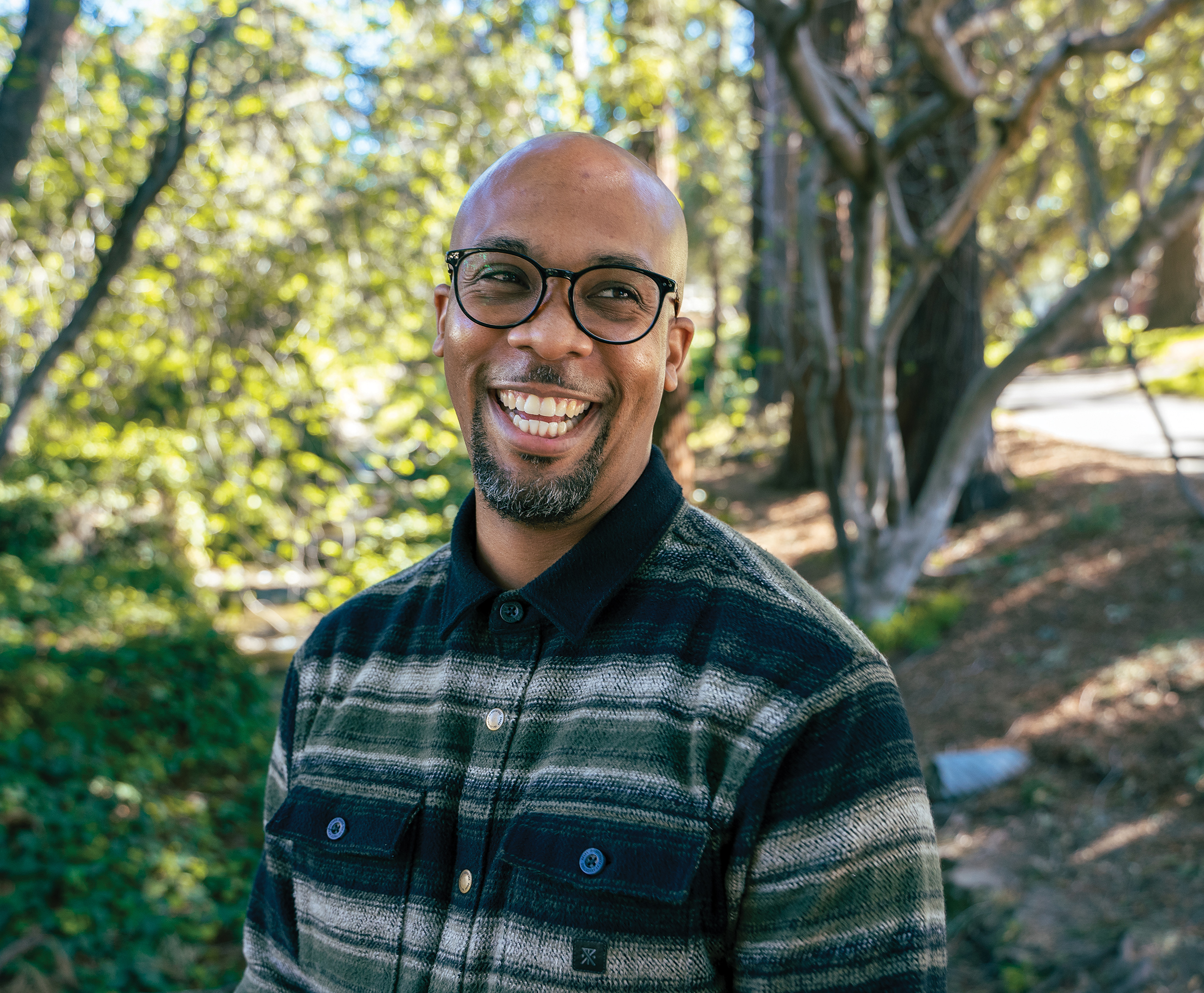 Photo of Kendall standing in a grove of trees, smiling and wearing a black/green/white striped shirt.