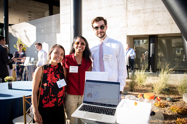 Color photo of three people standing around a laptop in the BBH courtyard, all smiling at the camera.