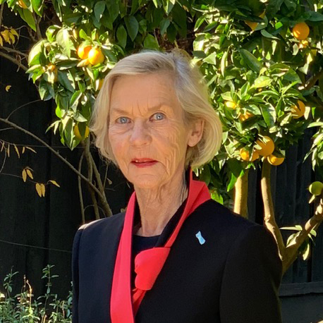 Color photo of Barbara with a blond bob and a red-lined black jacket, standing in front of a lemon tree.