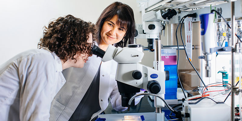 Color photo of a woman in a lab, smiling as another woman looks through a microscope