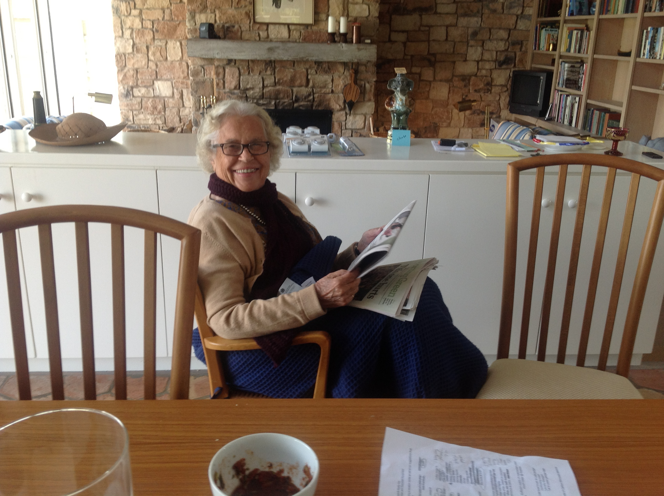 Photo of Becky smiling at her kitchen table with a paper in her hands and wrapped in a blue blanket.