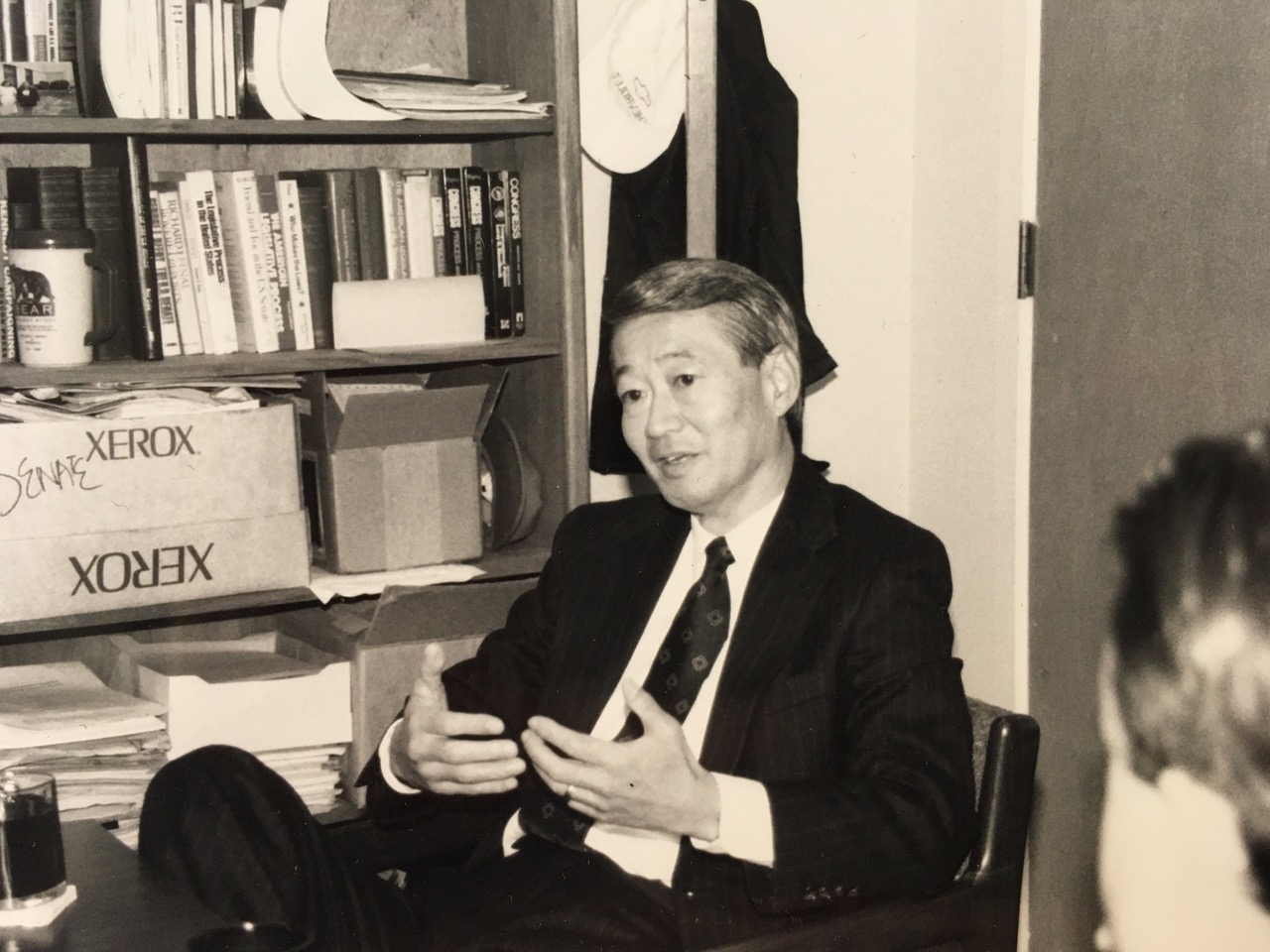 Black and white photo of Bob wearing a dark suit and sitting in a chair, with a crowded bookshelf behind him.