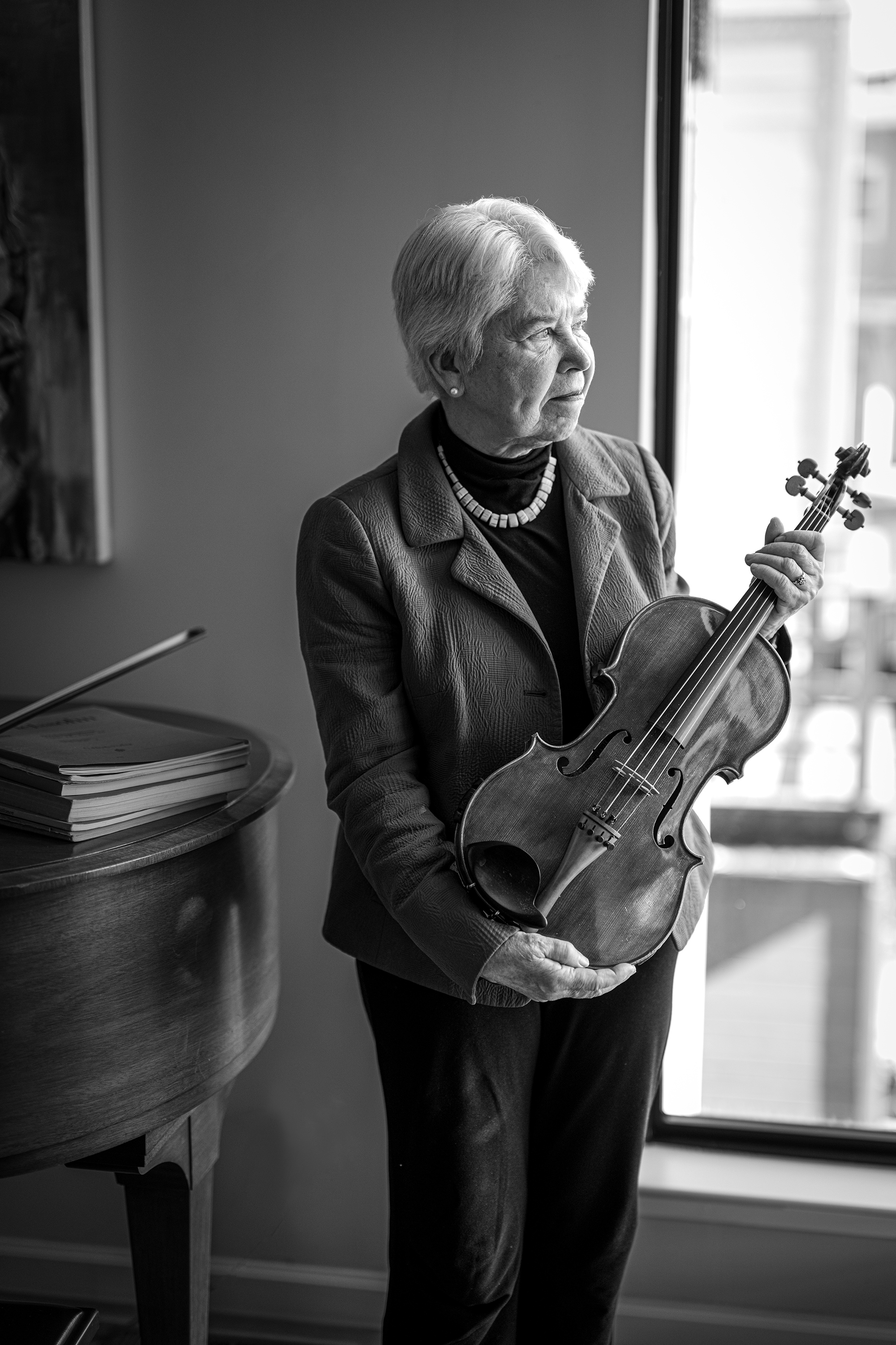 Black and white photo of the chancellor standing by a piano, holding her viola and looking out a window.