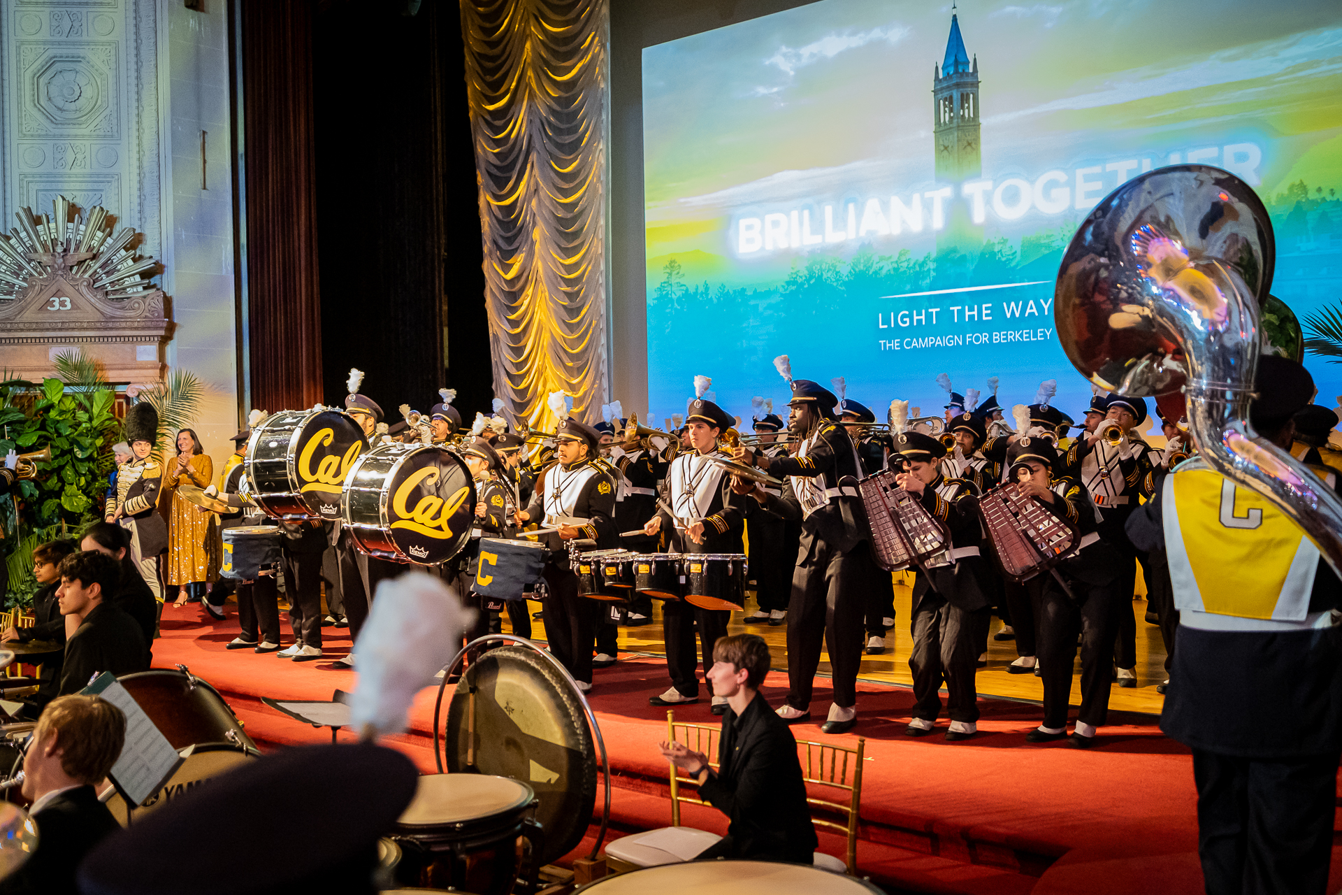 Photo of the band in uniform performing live in front of a large screen with a photo of the Campanile bathed in beautiful colors of yellow, green, and blue.