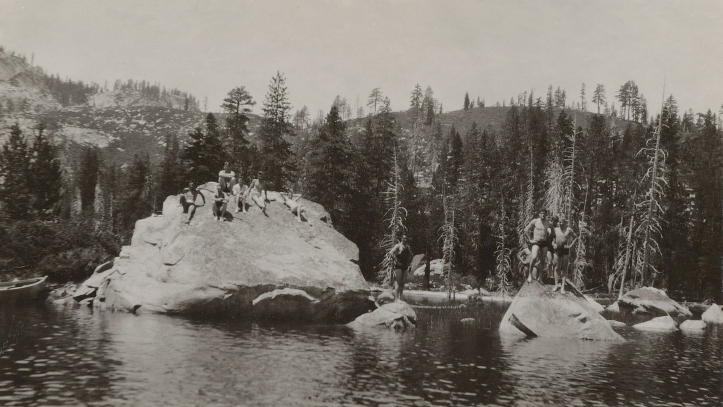 Black-and-white photo of eight people in swimsuits standing on two large rocks in a lake in front of tall trees with mountains in the distance