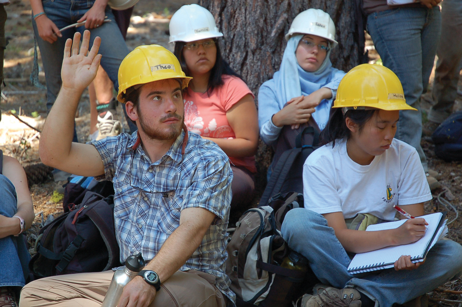 Photo of four students in hard hats sitting on the ground in front of a tree, one with hand raised and the rest listening intently